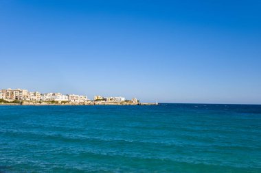 Berrak turkuaz deniz, modern beyaz binalar ve küçük bir liman fenerinin bulunduğu Otranto rıhtımına doğru uzanır. Kayalık kıyı şeridi canlı mavi bir gökyüzünün altındadır. Sahne, kıtır kıtır ufuk ve sakin deniz kenarı atmosferini ön plana çıkarıyor.