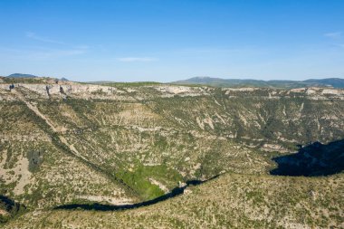 Geniş panoramik Cirque de Navacelles manzarası katmanlı kireçtaşı uçurumları, yeşil bitki örtüleri ve parlak mavi gökyüzünün altındaki derin gölgeler. Engebeli arazi ve engebeli yamaçlar dramatik bir doğal manzara yaratır..