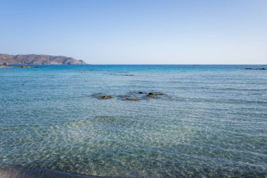 Ufuk boyunca uzanan sığ turkuaz su kayalık çıkıntılar ve geniş mavi gökyüzünün altında görünen yüzücülerle birlikte. Uzak tepeler kıyı şeridini çevreleyerek sakin ve davetkar bir sahil havası yaratıyor..