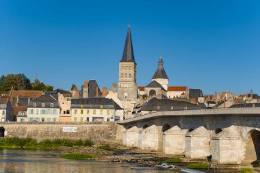Sivri uçlu manastır kulesi, Loire nehri boyunca uzanan klasik bir taş köprü ile La Charite sur Loire 'daki tarihi çatıların ve pastel evlerin üzerinde yükselir. Nehir kenarı sahnesi ortaçağ mimarisini ve huzur dolu cazibesini yakalıyor.