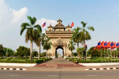 Laos 'un Vientiane kentindeki Patuxai anıtının merkezi manzarası, yüksek palmiye ağaçları ve ulusal bayraklar tarafından çerçevelenmiştir. Süslü yapı, açık gün ışığında manikürlü bahçelerin ortasında duruyor ve geleneksel Lao mimari ayrıntılarını vurguluyor..