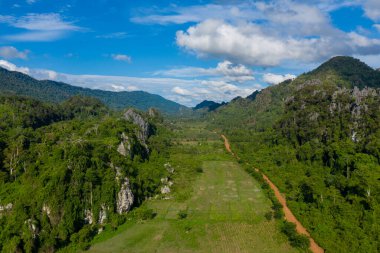 A winding red dirt road cuts through a lush rural valley bordered by dramatic limestone hills and dense tropical forest. The landscape is bathed in bright daylight, with scattered clouds and vibrant green vegetation under a vivid blue sky.