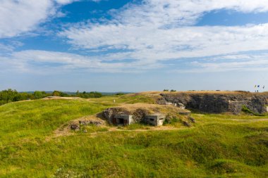 Eski taş sığınaklar, Fort de Douaumont 'taki çimenli, kraterli tepelerde dağınık bulutlu, parlak mavi bir gökyüzünün altında gömülüdür. Tarihi istihkam, ufukta görünen uzak bayraklarla açık yeşil manzaranın ortasında yer alıyor..