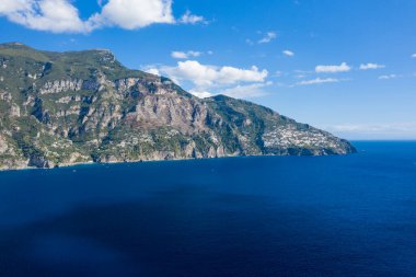 Amalfi Sahili boyunca sarp kayalıklarla ve yamaçlardaki köylerle buluşan derin mavi Akdeniz sularının geniş manzarası. Parlak güneş ışığı ve dağınık bulutlar canlı, sakin bir İtalyan kıyı manzarası yaratır..