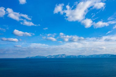 Uçsuz bucaksız mavi deniz dağınık beyaz bulutlarla dolu bir gökyüzünün altındaki uzak bir dağ sırasına doğru uzanıyor. Sakin sahne ince gradyanlar ve barışçıl, açık bir atmosfer içeriyor..