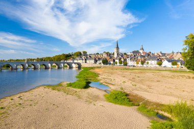 Ön plandaki geniş kumlu nehir kıyısı tarihi taş köprüye ve kilise kulesi ve parlak mavi gökyüzünün altındaki pastel evleriyle La Charite sur Loire 'ın ufuk çizgisine çıkar. İnce bulutlar ve gür yeşillik nehir kenarındaki cazibeyi vurgular.