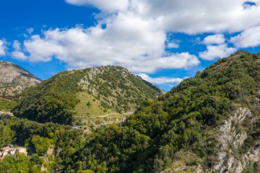 Tüylü beyaz bulutlarla dağılmış parlak mavi gökyüzünün altında yemyeşil tepeler dikleşir. Güneş ışığı, bu sakin kırsal bölgede yoğun yeşilliklerin ve kayaların çeşitli dokularını vurgular..