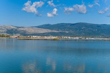 Yunanistan 'ın batısındaki sakin bir tuz gölü mavi gökyüzünü ve dağınık bulutları yansıtırken, Missolonghi kasabası ve uzak dağ yamaçları manzaralı bir zemin oluşturuyor. Sakin su ve geometrik kıyı şeridi açık alan ve doğal alan hissi yaratır.