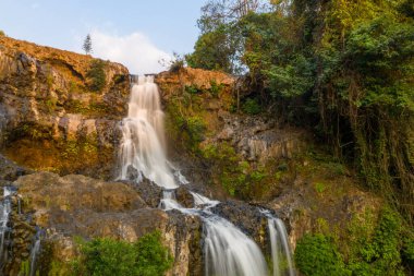 Sıcak güneş ışığı, Güney Laos 'taki Bolaven Platosu' ndaki yemyeşil tropikal bitki örtüsüyle çevrili engebeli bazalt kayaların üzerinden akan Tad Yuang şelalesinin çağlayan sularını vurgular. Kıvrımlı yamaç yüzeyi ve canlı yeşillik çarpıcı bir etki yaratır.