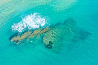İtalya 'nın Acciaroli kenti yakınlarındaki büyük bir su altı kaya oluşumunun üzerine düşen beyaz dalgalı temiz turkuaz suyun havadan görüntüsü. Güneş ışığı yüzeyin altındaki dokuları vurgular, canlı ve ferahlatıcı bir kıyı manzarası yaratır..