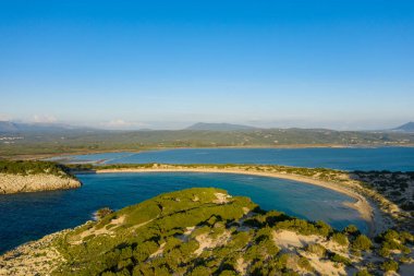 Bereketli yeşilliklerle çevrili hilal şeklinde kumlu bir sahil ve ufuktaki uzak dağlarla dolu canlı mavi gökyüzünün altında Paleokastro yakınlarında berrak turkuaz bir deniz..