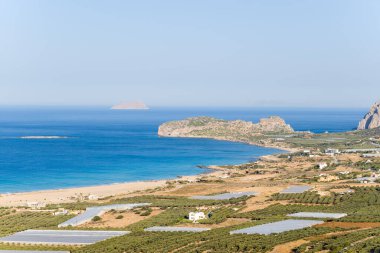 Girit 'teki Falasarna kıyı şeridini süpüren turkuaz deniz, kumlu kıyı şeridi, zeytinlikler ve açık mavi gökyüzü altında seralar yer alıyor. Sahne güneşli, ferah ve dünyevi Akdeniz tonlarıyla huzur dolu..