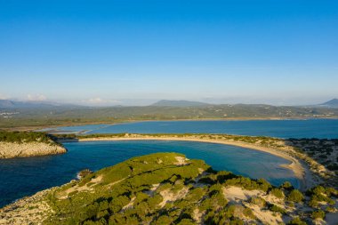 Parlak yeşil bitki örtüsüyle çevrili el değmemiş hilal şeklindeki kumsalın ve Paleokastro yakınlarında sakin mavi suyun günbatımında berrak bir gökyüzünün altında uzak dağların görüntüsü..