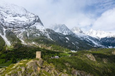 İtalya 'nın Val dAoste kentindeki yemyeşil Oyace vadisinde kayalık bir çıkıntının üzerinde ıssız antik bir taş kule duruyor. Karla kaplı dağ zirveleri ve yoğun ormanlar dinamik bir bahar gökyüzü altında dramatik bir zemin oluşturur..