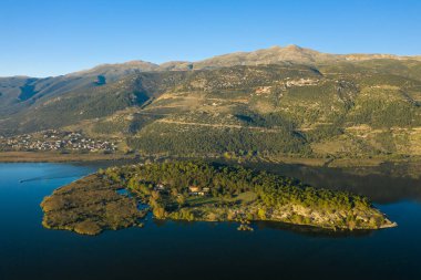 Ağaçlarla kaplı Ioannina Adası 'nın sakin mavi sularla çevrili havadan görünüşü, Pindus dağları temiz sabah ışığı altında arka planda yükseliyor. Sahne doğal dokuları, yemyeşil alanları ve huzurlu ortamı vurguluyor.