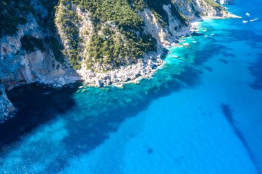 Bereketli yeşilliklerle kaplı engebeli uçurumların hava perspektifi Akdeniz 'in berrak, canlı mavi sularıyla Cala Goloritze, Baunei, Sardinya yakınlarında buluşuyor. Güneş ışığı kayalık dokuları ve turkuazın ve derin mavi denizin eğimini vurgular.