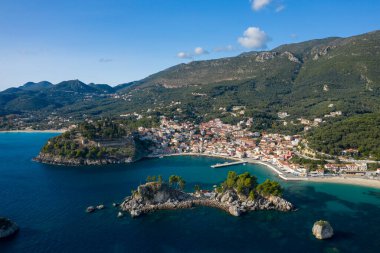Aerial perspective of Parga in Epirus, Greece, showcasing a vibrant coastal village, a historic hilltop fortress, and rocky islets surrounded by turquoise Ionian waters. The scene is framed by densely forested hills and distant mountains under clear