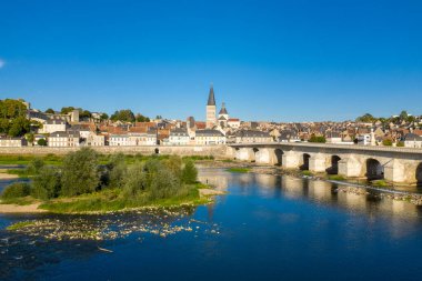 Loire nehri boyunca uzanan tarihi taş köprüsü, geleneksel evleri ve canlı mavi gökyüzünün altındaki kilise kulesiyle La Charite sur Loire 'ın geniş açılı manzarası. Nehir kıyısı boyunca yemyeşil ve durgun sudaki berrak yansımalar huzur uyandırıyor.