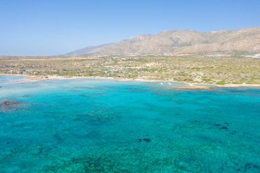 Girit 'teki Elafonisi plajının geniş hava perspektifi, berrak turkuaz deniz, kumlu sahil şeridi ve bulutsuz mavi gökyüzünün altındaki engebeli tepeler sergileniyor. Canlı renkler ve açık arazi özgürlük ve doğal güzelliği çağrıştırıyor..