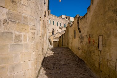 Matera, İtalya 'da temiz mavi gökyüzünün altında yıpranmış kireçtaşı duvarlarla çevrili boş kaldırım taşlı bir sokak. Sıcak güneş ışığı sert dokuları vurgular ve zamansız Akdeniz hissi için güçlü gölgeler yaratır..