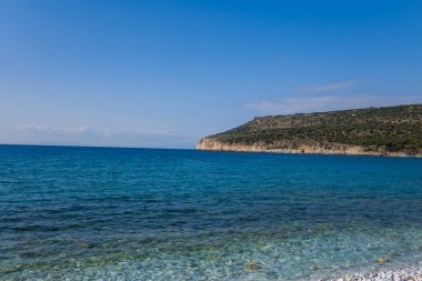 Şeffaf mavi suyu olan parlak bir çakıl taşı sahili ve berrak bir gökyüzünün altında yeşil bitki örtüsüyle kaplı engebeli bir burnu. Güneş ışığı, Akdeniz kıyı şeridinin canlı renklerini ve doğal dokularını güçlendirir..