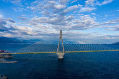 Yunanistan 'ın batısındaki Patras yakınlarındaki Rion-Antirion Köprüsü, çarpıcı asma kabloları ve gökyüzünün altında dokulu bulutlarla dolu bir dağ zeminiyle derin mavi suları kaplıyor. Modern yapı doğal manzaraya karşı duruyor.