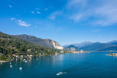 Maggiore Gölü 'nün geniş bir manzarası. Derin mavi su, ormanlık yamaçlar ve berrak bir gökyüzünün altındaki engebeli dağlar. Küçük bir tekne gölde hafif dalgalanmalar bırakırken, göl kenarındaki köyler kuzey İtalya 'da kıyı şeridi boyunca yuva yapıyor..