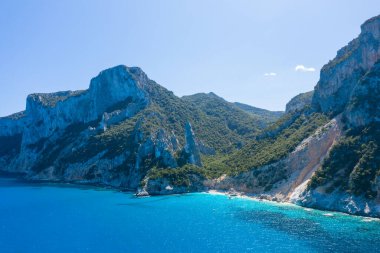 Cala Goloritze, Baunei, Sardinya yakınlarındaki canlı turkuaz bir körfezin üzerinde yemyeşil yamaçlar ve dramatik kireçtaşı uçurumlar yükseliyor. Parlak güneş ışığı ve açık mavi gökyüzü engebeli Akdeniz kıyı şeridini vurguluyor.