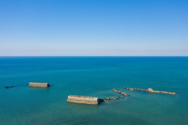 Asnelles, Normandiya açıklarında engin bulutsuz bir gökyüzünün altında açık mavi-yeşil sulara dağılmış yapay bir limandan geniş beton bloklar ve parçalanmış yapılar..