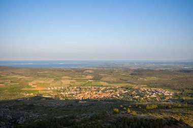 Expansive view over a rural village surrounded by patchwork fields and rolling hills, with the shimmering coastline visible on the horizon under a clear blue sky near Opoul Perillos.