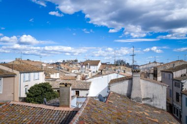 Aigues-Mortes 'in tarihi merkezinde geleneksel kiremitli çatılara ve kırsal cephelere dramatik bulutlar ve açık mavi gökyüzü ile yükseltilmiş perspektif. Sahne, güney Fransız mimarisinin cazibesini vurgulayarak havadar ve dokulu hissettiriyor.