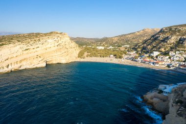 Girit 'teki Matala Körfezi' ne geniş bir hava perspektifi. Geniş çizgili kireçtaşı kayalıkları, derin mavi su ve temiz güneş ışığı altındaki küçük bir sahil köyüyle çevrili kumlu bir sahil..