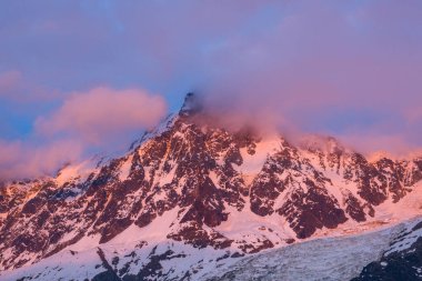 Sıcak pembe ve mor ışık Aiguille du Midi 'nin engebeli, karla kaplı zirvesinde Fransız Alpleri' nde alacakaranlıkta zirveye yumuşak bulutlar sürükleniyor..