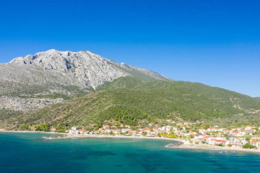 Kırmızı çatılı evleri olan güneşli bir sahil köyü, turkuvaz suları ve berrak mavi gökyüzünün altındaki yemyeşil tepelerle çevrili dramatik, kayalık bir dağın dibinde yer alır. Huzurlu Akdeniz manzarası tazelik ve doğal cazibe yayar..