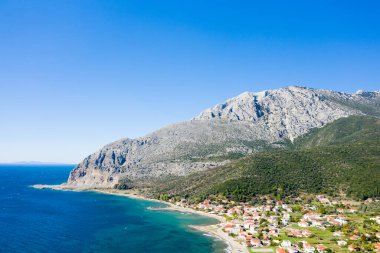 Geniş bir hava perspektifi Kato Vasiliki köyünü, yoğun yeşillik ve açık mavi gökyüzü ile dramatik bir kayalık dağın altındaki turkuaz bir kıyı şeridi boyunca yakalıyor..