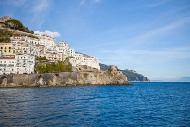 Pastel ve beyaz boyalı binalar İtalya 'nın Amalfi kentindeki sarp kayalıklar boyunca yığılmıştır ve derin mavi Akdeniz manzarası vardır. Tarihi taş kemerler ve kıyı gözetleme kulesi açık gökyüzünün altındaki dramatik kıyı şeridini vurguluyor..