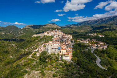 İtalya 'nın Rivello kentinin hava perspektifi, canlı mavi gökyüzü altında dramatik dağlarla çevrili yemyeşil bir yamaçtan dökülen terakotta çatıları olan tarihi bir tepe köyünü gözler önüne seriyor. Kıtır gün ışığı ve dağınık bulutlar canlı bulutları güçlendirir