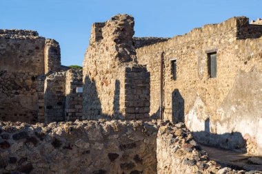 Yıpranmış volkanik taş ve tuğla duvarlar İtalya 'nın Pompeii kentindeki eski evlerin kalıntılarını oluşturuyor. Sıcak güneş ışığı, açık mavi gökyüzünün altındaki kaba dokuları ve geometrik çizgileri vurgulayan güçlü gölgeler oluşturur..