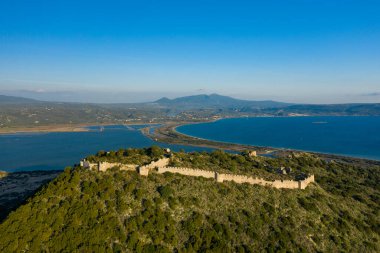 Navarino Körfezi 'nin süpürücü mavi sularının üzerinde yeşil ormanlı bir tepeyi taçlandıran antik taş kale duvarlarının hava görüntüsü, berrak bir gökyüzü altında uzak dağlar..