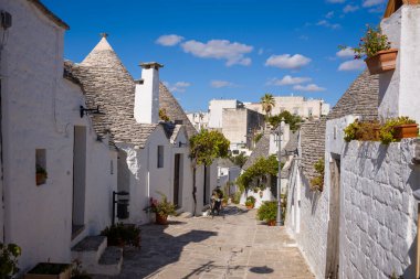 Konik taştan çatıları olan geleneksel trullo evleri İtalya 'nın Apulia eyaletinin Alberobello şehrinde güneşli dar bir caddeye sahiptir. Saksı bitkileri, desenli duvarcılık ve canlı mavi bir gökyüzü kendine özgü bir güney İtalyan köyü atmosferi yaratır..