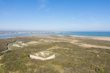 Yoğun kıyı bitkileri ve kumlu kumullarla çevrili taştan bir kalenin havadan görünüşü. Geniş mavi deniz ve açık bir gökyüzünün altındaki uzak kıyı şeridi..