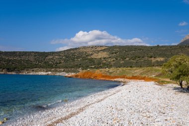 Temiz mavi denizle buluşan el değmemiş, beyaz bir çakıl taşının geniş manzarası. Çarpıcı turuncu uçurumlar ve parlak bir gökyüzünün altındaki yeşil tepeler. Güneş ışığı, canlı kıyı renklerini ve doğal dokularını güçlendirir..