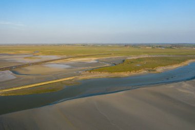 Geniş gel-git bataklıkları ve açık mavi gökyüzünün altında yeşil çayırlarla çevrili dolambaçlı su kanallarının hava manzarası. Açık arazi yumuşak ışık, ince dokular ve sakin bir atmosfere sahiptir..