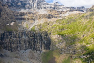 Dik taş duvarlar ve Gavarnie Sirki 'ndeki çimenli yamaçlar boyunca aydınlık ve gölge oynar. Parçalı bulutlu bir gökyüzünün altında engebeli dokuları ve ince dereleri aydınlatır..