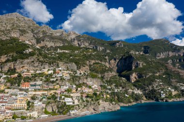 Parlak güneş ışığı, İtalya 'nın Positano kentindeki renkli tepe binalarını aydınlatıyor. Dik kireçtaşı uçurumlarına ve parlak mavi gökyüzüne karşı. İkonik Amalfi Sahili manzarası dramatik suların altında yemyeşil bitki örtüsü ve turkuaz Akdeniz suları barındırıyor.