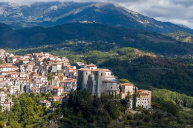 İtalya 'nın Rivello kentinin manzarası yükseldi. Toplu teraslarla çevrili tarihi bir taştan kiliseyi ön plana çıkarıyor. Yeşillik, yeşil orman ve bulutlu bir gökyüzünün altında dramatik dağ yamaçları var..