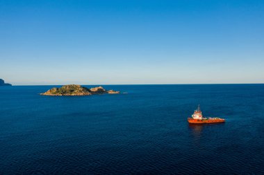 Canlı kırmızı bir çalışma teknesi, kayalık yeşil Isola dellOgliastra yakınlarında açık gökyüzü ve Sardunya 'nın Lotzorai kenti yakınlarındaki uzak kıyı şeridi ile derin mavi Akdeniz sularında yüzer. Sahne, sakin denizciliği vurgulayarak, canlı gün ışığında yıkanır.