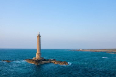 Cap de la Hague 'deki turkuaz denizle çevrili kayalık bir adadan, açık mavi gökyüzü ve arka planda uzak bir kıyı şeridi bulunan yalnız bir deniz feneri yükseliyor. Olay yeri gün ışığıyla yıkanmış, deniz kuvvetini çağrıştırıyor ve...