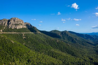 Sardinya, Baunei ve Dorgali yakınlarındaki yoğun yeşil ormanlar ve dramatik uçurumlar boyunca yükselen yol. Manzara güneşli, geniş ve engebeli bir gökyüzü altında engebeli tepelerle çevrili..