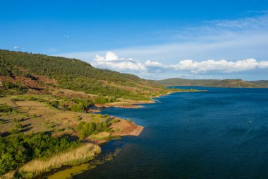 Salagou Gölü 'nün engin manzarası. Derin mavi su, yeşil tepeler, kırmızı toprak ve bulutlarla kaplı parlak gökyüzü altında uzun kamışlarla buluşuyor..